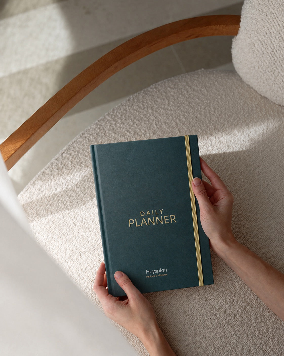 Twee handen houden de donkergroene HuysPlan daily planner vast boven een boucle stoel